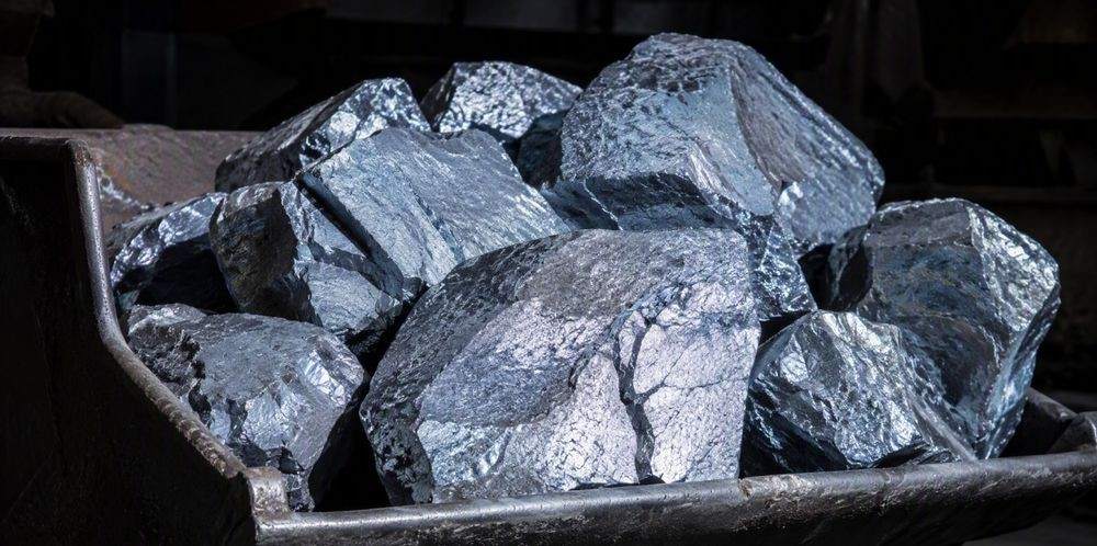 A heap of silver-gray silicon metal lumps with bright crystalline fracture faces piled in a steel scoop.
